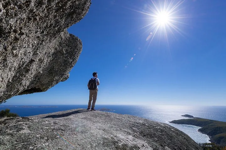 Person with backpack standing on a large rock overlooking the ocean under bright sun.