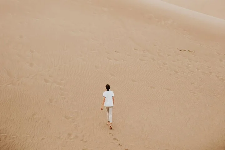 Person walking barefoot across extensive sand dunes leaving a trail of footprints.